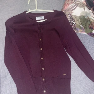 Vinröd kofta från Holly & Whyte - Snygg vinröd kofta från Holly & Whyte med rund halsringning och guldiga knappar framtill. Långärmad och i mjukt, tunt stickat material som passar perfekt till lager-på-lager. Enkel och stilren design som funkar till många outfits. Använd 1 gång så i super skick, storlek xs!