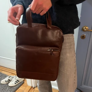 Brun skinn portfölj med dragkedja - Snygg portfölj i mörkbrunt skinn med stilren, fyrkantig form. Väskan har två rejäla handtag, flera fack med dragkedja både fram och bak samt ett rymligt huvudfack. Perfekt för laptop och viktiga papper. Klassisk och tidlös design som passar till allt. I äkta läder! Helt ny aldrig använd. Ny 1795kr