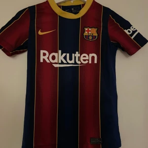 FC Barcelona matchtröja Nike - Säljer en officiell FC Barcelona matchtröja från Nike med Rakuten-tryck. Tröjan har klassiska blå och röda vertikala ränder med gula detaljer och klubbmärke på bröstet. Kortärmad modell i lätt och ventilerande material, perfekt för fotboll eller supporterliv.