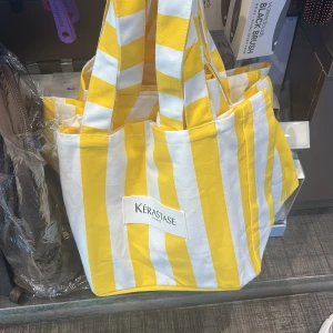 Randig shoppingväska från Kérastase - Gul och vit randig shoppingväska från Kérastase med breda axelband och rymlig form. Väskan är tillverkad i bomull och har en tydlig logotyp framtill. Perfekt för att bära med sig allt man behöver under dagen.