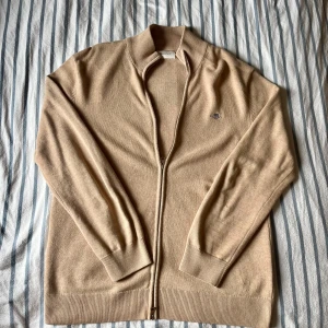 Beige kofta med dragkedja från GANT - Säljer en stilren beige kofta från GANT med hel dragkedja och broderad logga på bröstet. Tröjan är ribbstickad vid krage, ärmslut och nederkant och är gjord i mjuk bomull. Perfekt för lager-på-lager och snygg till jeans eller chinos.