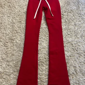 Röda bootcut mjukisbyxor Hidden Cult XS - Röda mjukisbyxor från Hidden Cult med vit ribbad midja och snörning. Bootcut-modell med broderad vit logga bak. Sköna och snygga byxor i mjukt material, perfekta för en chill och trendig look.