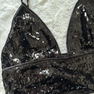 Svart paljettopp från NA-KD - Säljer en svart glittrig paljettopp från NA-KD med triangelformade kupor och smala axelband som korsas i ryggen. Toppen har en croppad passform och dragkedja i sidan. Perfekt för att sticka ut med extra bling.