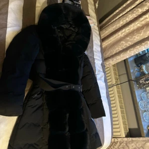 Svart pufferjacka med fuskpäls Copperose - Säljer en svart pufferjacka från Copperose i storlek M. Jackan har en stor huva och breda detaljer i fuskpäls längs huva och framkant. Midjebälte med spänne ger snygg siluett. Perfekt för kalla dagar när du vill vara både varm och stilren.