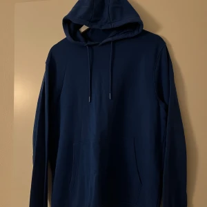 Mörkblå hoodie från Dressmann - Säljer en mörkblå hoodie från Dressmann i storlek L. Tröjan har huva med snörning och en stor magficka framtill. Tillverkad i bomull.