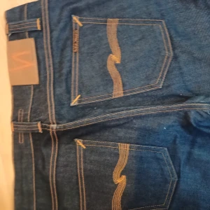 Mörkblå jeans från Nudie Jeans, 28/32 - Snygga mörkblå jeans från Nudie Jeans med klassisk orange söm och ikonisk bakficka. Modellen har normal passform och raka ben. Tillverkade i slitstark denim av bomull. Perfekta för dig som gillar stilrena och tidlösa jeans.