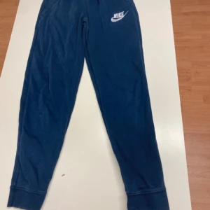 Blå mjukisbyxor från Nike - Mörkblå mjukisbyxor från Nike med resår och snörning i midjan. Byxorna har ribbade muddar vid bensluten och klassisk Nike-logga broderad på ena benet. Perfekta för chill eller träning, riktigt sköna och stilrena.