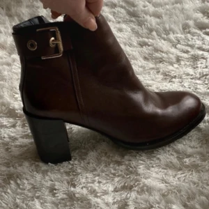 Bruna stövletter från Tommy Hilfiger - Snygga bruna stövletter från Tommy Hilfiger med kraftig klack och dekorativt spänne i guld vid ankeln. Skorna är tillverkade i skinn och har en rund tå samt platt sula med mönstrad undersida. Perfekt för dig som gillar stilrena boots med lite edge.