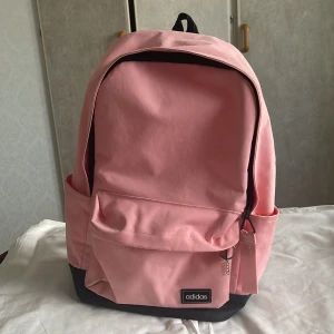 Rosa ryggsäck från Adidas - Snygg rosa ryggsäck från Adidas med svart botten och detaljer. Väskan har ett stort huvudfack, framficka med dragkedja och två sidofickor. Justerbara axelremmar och klassisk Adidas-logga framtill. Perfekt för skola eller cityhäng.