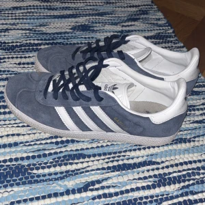Adidas Gazelle sneakers blå/vit - Säljer ett par klassiska Adidas Gazelle sneakers i mörkblå mocka med vita detaljer och de ikoniska tre ränderna på sidan. Skorna har snörning, platt sula och rund tå. Adidas-logga på häl och plös. Perfekta för dig som gillar retrostil och streetwear.
