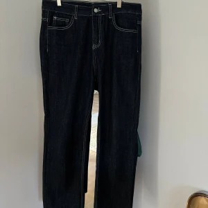 Mörka denimjeans med kontrastsömmar – raka ben - Välsydda jeans i mörk tvätt med tydliga vita kontrastsömmar och klassisk 5-ficksdesign. Tidlös rak passform som fungerar till både vardags och mer dressade tillfällen. Jeansen är oanvända och i nyskick. Passar dig som gillar rena linjer med subtila detaljer.  • Midja: 84 cm • Höft: 114 cm • Benlängd: 103 cm • Fotvidd: 44 cm  Storlek: Passar dig som bär W32 beroende på fit