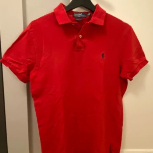 Röd pikétröja från Ralph Lauren - Snygg röd pikétröja från Polo Ralph Lauren i custom fit. Klassisk krage, korta ärmar och blå broderad logga på bröstet. Tillverkad i mjuk bomull som är skön mot huden. Perfekt för en stilren och sportig look.