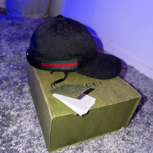 Svart Gucci keps med GG-mönster - Snygg svart keps från Gucci med klassiskt GG-monogram över hela kepsen. Kepsen har en grön och röd rand på sidan och justerbar rem baktill i skinn. Tillverkad i bomull och detaljer i skinn. Perfekt för dig som vill ha en lyxig och stilren accessoar.