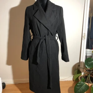 Svart kappa från Bik Bok i ullmix - Lång svart kappa från Bik Bok i ullblandning. Klassisk modell med bred krage, knytskärp i midjan och stora fickor framtill. Fickorna har tyvärr hål inuti. Ärmlängd ca 65cm. Längd ca 115cm. Pris: 80kr