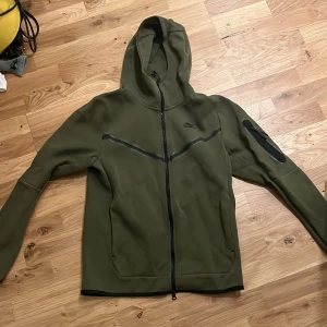 Mörkgrön Nike Tech Hoodie - Nike hoodiejacka i olivgrönt med hel dragkedja framtill och huva. Jackan har svarta detaljer, sned bröstpanel och ficka med dragkedja på ena ärmen.