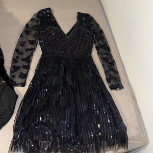 Svart glittrig kortklänning med paljetter - Supersnygg svart kortklänning med v-ringning, långa transparenta ärmar och massor av glittrande paljetter. Ärmarna har dekorativa mönster och klänningen har en markerad midja samt ett skirt, flowy tyg. Perfekt för dig som vill sticka ut med en unik look! Köpt för 2000