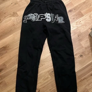 Trapstar Byxor med Puff Print - Svarta mjukisbyxor från Trapstar med stort broderat Trapstar-logo i grått och silver framtill. Byxorna har elastisk midja med snörning och raka ben.