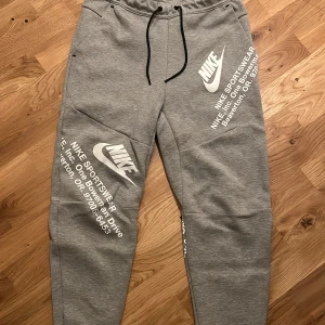 Grå & Vita Nike Tech Byxor - Snygga grå mjukisbyxor från Nike med vit logga och texttryck på benen. Byxorna har svarta snören i midjan, dragkedja på fickan och en tapered passform.