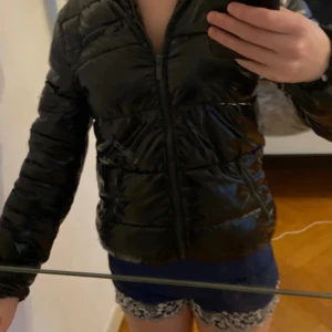 Svart glansig pufferjacka Fishbone XS - Säljer en svart glansig pufferjacka från Fishbone i storlek XS. Jackan har huva, dragkedja framtill och två fickor med dragkedja. Den är quiltad och har en riktigt snygg streetstil. Perfekt för dig som vill ha en varm och trendig jacka med lite extra attityd.