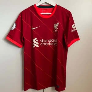 Liverpool hemmatröja 2021/2022 - Liverpool hemmatröja från säsongen 2021/2022 med Mohammed Salah på ryggen. Även Premier League loggan finns på höger ärm. Köpt på unisport för 1399kr, mitt pris 250kr. Helt oanvänd, bara hängt i garderoben. Hör av er vid frågor & funderingar!