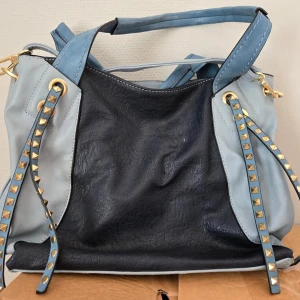 Blå axelväska med nitar - Snygg axelväska i blått skinn med coola guldfärgade nitar på remmarna. Väskan har flera fack med dragkedja och breda axelremmar i ljusblått. Perfekt för dig som vill ha en unik och rymlig väska med detaljer som sticker ut.