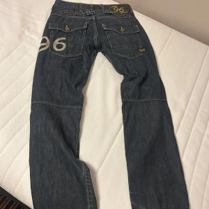 Sällsynt modell G star jeans storlek 31/32 - Säljer ett par mörkblå jeans från G-Star med coolt 96-tryck på bakfickan och benet. Byxorna har stora fickor med lock och knappar bak, samt detaljerade sömmar över knäna för en schysst streetstil. Passar dig som gillar unika jeans med lite edge.