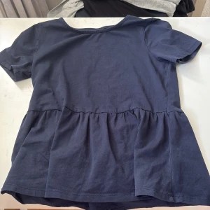 Mörkblå blus från Lindex Kids - Supersöt mörkblå blus från Lindex Kids i mjuk bomull. Modellen har korta ärmar och en volang nedtill som ger en avslappnad och trendig look. Perfekt för dig som gillar enkel men snygg stil.