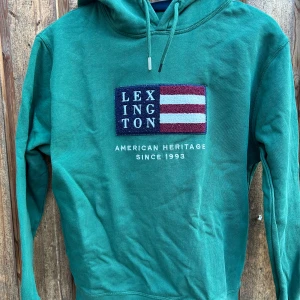 Grön hoodie från Lexington - Snygg grön hoodie från Lexington med broderad flagga och text på bröstet. Tröjan har huva med snörning och är tillverkad i mjuk ekologisk bomull. Perfekt för dig som gillar klassisk och avslappnad stil.Storlek 158/164