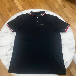 Svart Prada pikétröja med röd/vit kant - Svart pikétröja från Prada med korta ärmar och klassisk krage. Snygga röda och vita ränder på krage och ärmslut ger en sportig vibe. Tröjan har knappar framtill och diskret Prada-logga på bröstet. Materialet är bomull och modellen har en normal passform. nypris 530 euro. 