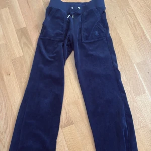 Mörkblå mjukisbyxor Juicy Couture XXS - Snygga mörkblå mjukisbyxor från Juicy Couture i storlek XXS. Byxorna har bred resårmidja med snörning, stora fickor fram och bak med knappar, och är gjorda i mjuk sammet. Perfekta för chill dagar och har en klassisk straight passform.