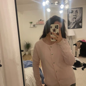 Ljusrosa kofta med silverknappar - Supersöt ljusrosa kofta med rund halsringning och stilrena silverfärgade knappar framtill. Koftan har lång ärm och en clean, enkel look som passar perfekt till lager-på-lager. Mjuk och skön, troligtvis i bomull eller syntet.