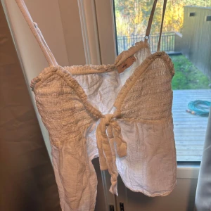 Vit linnetopp med knyt  - Supersöt vit linnetopp från Nelly med smala axelband och snygg knytning framtill. Toppen har smockad överdel och är tillverkad i ett lätt, krispigt material som känns perfekt för varma dagar. Passar dig som gillar en chill och feminin stil.
