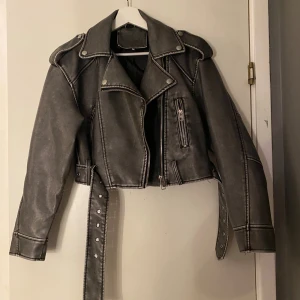 Grå croppad skinnjacka med bälte - Snygg grå croppad skinnjacka med coola detaljer som breda axlar, silverfärgade knappar och dragkedjor samt ett långt bälte med nitar. Jackan har klassisk bikerstil och är perfekt för dig som vill ha en edgy look. Passar dig som gillar street och rock vibes.❤️