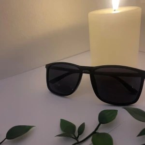 Svarta solglasögon med fyrkantig form - Snygga svarta solglasögon med fyrkantig form och mörka glas. Skalmarna har en diskret textur och en liten detalj i rött och grönt. Perfekta för att ge en cool vibe till din outfit.