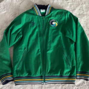 Grön bomberjacka från Umbro - Snygg grön bomberjacka från Umbro med broderad logga och New York Cosmos-märke på bröstet. Jackan har dragkedja framtill, randiga muddar i blått, gult och vitt samt två fickor med dragkedja. Perfekt för dig som gillar sportig stil.