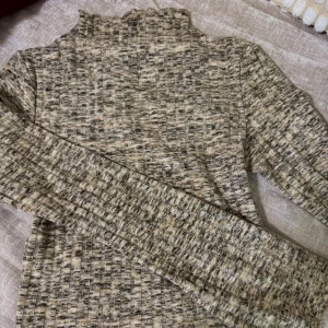 Rutig polotröja från BikBok XS - Säljer en beige och gråmelerad rutig polotröja från BikBok i storlek XS. Tröjan har lång ärm, ribbad struktur och hög hals med vågig kant. Perfekt för lager-på-lager och snygg till jeans eller kjol.