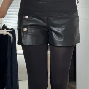 Svarta skinnshorts med guldknappar - Snygga svarta shorts i skinnimitation med två dekorativa guldknappar på sidan. De har en rak passform och är lite kortare i modellen, vilket ger en cool och trendig vibe. Perfekta att styla med tights eller bara ben för en edgy look.