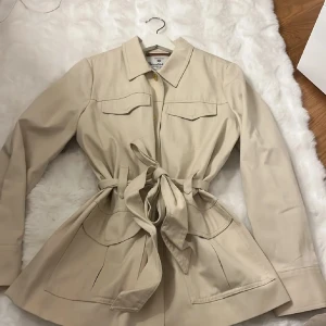 Beige overshirt från Bondelid med knyt - Snygg beige overshirt från Bondelid med klassisk krage, stora fickor och knyt i midjan. Perfekt för dig som gillar stilrena och moderna plagg. Jackan har långa ärmar och knappar framtill. Passar dig som vill ha en chill och clean look.