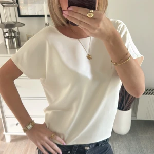 Vit omlottopp med knyt från Zara - Säljer en vit omlottopp från Zara i storlek S. Toppen har korta ärmar, rund hals och snygg öppen rygg med knyt bak. Materialet är mjukt och har en lätt glansig finish som ger en lyxig känsla. Perfekt att styla med jeans eller kjol för en trendig look.