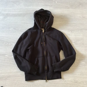 Svart hoodie från Burberry med dragkedja - Svart hoodie från Burberry med guldfärgad dragkedja, snörning i huvan och broderad logga på bröstet. Insidan av huvan har klassiskt rutigt Burberry-mönster. Mjuk bomullskänsla och känguruficka framtill. Perfekt för en stilren och chill look.