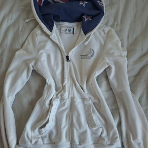 Vit hoodie Emirates Team NZ - Jag säljer denna jättefina new zeland zip upen med coolt mönster på luvan eftersom att den tyvärr inte kommer till användning. Den har några små fläckar som ni kan se i bilderna, men jag ska tvätta det en gång till innan jag skickar det till er. Man ser även inte fläckarna på långt håll. Och 3 små hål som ska gå att sy om. Priset kan diskuteras och skriv om ni vill ha fler bilder eller om ni undrar något om priset🥰.