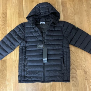 Svart dunjacka från Stone Island - Svart dunjacka från Stone Island med huva och klassisk logotyp på ärmen. Jackan är quiltad med horisontella sömmar, har dragkedja framtill och två sidofickor. Perfekt för kyliga dagar och har en clean, modern look.