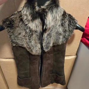 Brun pälsväst från AmoreSS Stockholm - Säljer en snygg brun pälsväst från AmoreSS Stockholm i storlek XXS. Västen har en fluffig krage och axelparti i brunmelerad päls, med kropp i mjuk mocka. Dragkedja framtill och två fickor med lock. Perfekt för att ge din outfit extra edge.