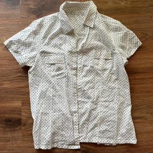 Polka dot blus  - Snygg vit skjorta från Cappuccini med svarta prickar, korta ärmar och klassisk krage. Skjortan har två bröstfickor med knappar och är tillverkad i mjuk bomull. Perfekt för dig som gillar en lekfull men stilren look.