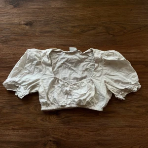 Vit topp med puffärmar - Säljer en vit croppad blus från Hess Frackmann med puffiga korta ärmar och elastisk kant vid midjan och ärmslut. Blusen har en söt spetsdetalj runt halsringningen och en lätt rynkad front. Perfekt för dig som gillar romantisk och vintage-inspirerad stil.