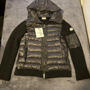 Svart pufferjacka från Moncler - Snygg svart pufferjacka från Moncler med glansig quiltad front och huva. Ärmarna är stickade och har en liten ficka med dragkedja på vänster arm. Jackan har två fickor framtill och stängs med dragkedja. Har använt jackan 2-3 gånger och är perfekt för kyliga dagar.