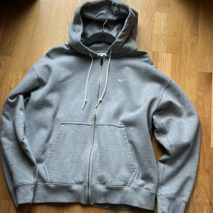 Grå hoodie med dragkedja från Nike - Grå zip-hoodie från Nike med liten vit logga på bröstet. Mjuk fleece på insidan, huva med snörning och två stora fickor framtill. Perfekt för chill eller när du vill ha något bekvämt och stilrent. Klassisk passform och långärmad.