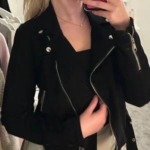 Svart bikerjacka med dragkedjor - Snygg svart bikerjacka med silvriga dragkedjor och knappar. Jackan har klassisk krage, bälte i midjan och detaljerade sömmar. Materialet är mjukt och matt, vilket ger en cool look. 