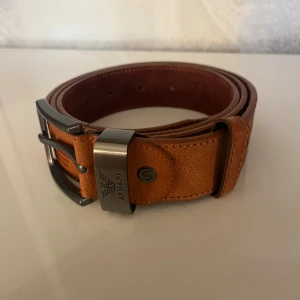 Brunt skärp från Armani i skinn - Snyggt brunt bälte från Armani i äkta skinn med präglad logga och metallspänne. Bältet har en klassisk form med stilrena sömmar och detaljer i silverfärgad metall. Perfekt för dig som vill ha en clean och exklusiv look.