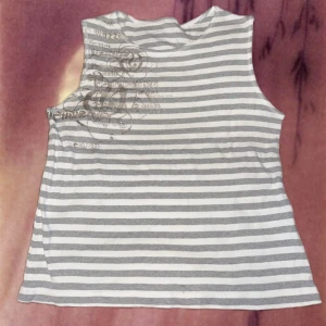Randig gråvit ärmlös linnetopp - Säljer en ärmlös linnetopp med grå och vita horisontella ränder. Toppen har ett diskret tryck med silvriga detaljer på ena axeln.